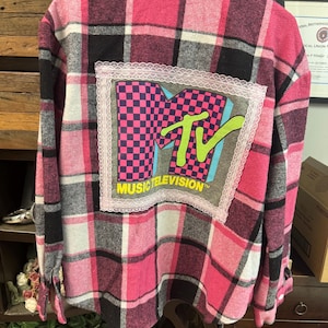 May include: A long-sleeve button-down shirt in a pink, black, and white plaid pattern. The back features a rectangular patch with the MTV logo in a pink and black checkerboard print, a bright green TV graphic, and the text MUSIC TELEVISION.