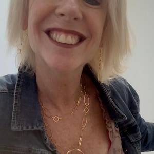 May include: A gold-toned necklace with a large, irregular circular pendant and a chain of connected oval and circular links. Matching gold earrings hang from the ears. The person is wearing a denim jacket.