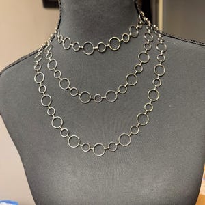 May include: Three silver-toned necklaces with a linked circle design are displayed on a black mannequin. The necklaces vary in length, with the shortest at the top and the longest at the bottom. The circles are uniform in size and evenly spaced.