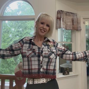 May include: A plaid shirt in shades of red, white, and blue, with a floral embellishment. The shirt has a cropped design and is paired with a white belt with silver studs. The shirt is a casual style.