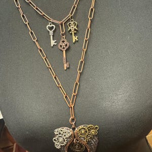 May include: A steampunk-style necklace with a copper chain and multiple key charms. The necklace features a large heart-shaped pendant with gears and a keyhole design. The keys are silver, gold, and copper-toned.