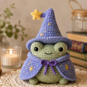 May include: A crocheted frog wearing a purple wizard hat and cape, adorned with yellow stars. The frog has large black eyes and a small smile. A yellow star is attached to the hat. The frog is sitting on a cream-colored doily.