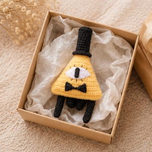 May include: A yellow, black, and white crochet triangle figure with a top hat, bow tie, and one eye, presented in a brown gift box with white tissue paper. The figure has black legs and is likely a character from a cartoon.