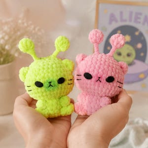 May include: Two handmade crochet alien plush toys. One is lime green, the other is pink. Both have black eyes, whiskers, and antennae with small pink or green balls on top. The toys are held in a person's hands.