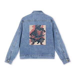 以下が含まれることがあります： 背中にカラフルなグラフィックが施されたライトブルーのデニムジャケット。グラフィックは、ピンクのアクセントが付いた赤と黒の悪魔のような姿を描いています。ジャケットは、ボタン留めとポケットを備えたクラシックなデザインです。