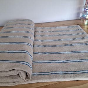 May include: A folded piece of beige fabric featuring horizontal stripes in white and dark blue. The textile has a visible woven texture and is laid out flat, with one section rolled over itself to display the pattern across the entire surface.