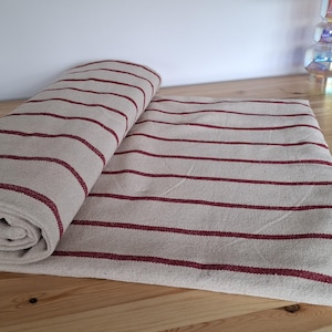 May include: A roll of fabric featuring a natural beige base color with horizontal dark red stripes. The textile is partially unrolled, displaying a consistent striped pattern across the surface of the material, which has a visible woven texture.