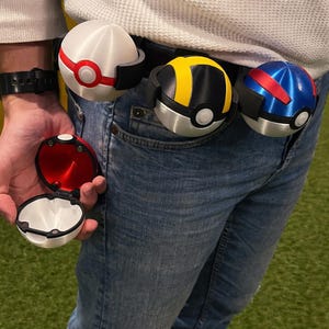 Könnte beinhalten: Eine Person trägt eine blaue Jeans und einen schwarzen Gürtel mit drei farbenfrohen Pokéball-Accessoires. Einer ist geöffnet und zeigt einen roten Innenraum. Die anderen drei sind am Gürtel befestigt und haben rote, gelbe und blaue Designs.