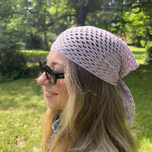 May include: A light lavender crochet headscarf featuring an openwork triangular pattern with delicate scalloped edges. The headscarf is tied at the back of the head with two thin, matching crocheted fabric ties, creating a soft and textured appearance.