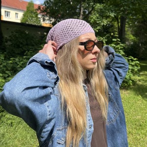 May include: A light purple crochet head covering featuring an openwork stitch pattern. The head covering is constructed from yarn and fits snugly over the top of the head, displaying a textured, grid-like appearance throughout the fabric.