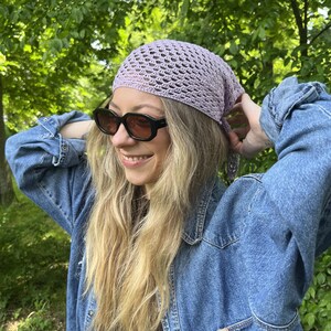 May include: A light purple crochet headscarf with a triangular shape and an openwork pattern, worn tied at the back of the head. The headscarf is paired with black-framed sunglasses and a blue denim jacket with long sleeves and a buttoned front.