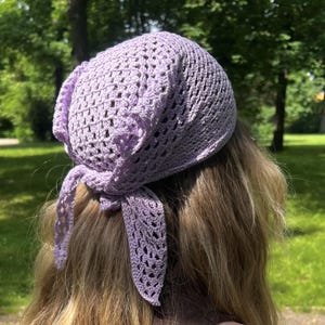 May include: A light purple crochet headscarf featuring an openwork, triangular pattern with a delicate, lacy texture. The headscarf is tied at the back with two pointed ends, creating a secure fit while showcasing the intricate, hand-stitched yarn design.