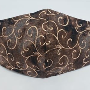 May include: A brown face mask with a gold swirling pattern. The mask has black ear loops and is made of fabric. The design is a repeating pattern of gold swirls on a brown background.