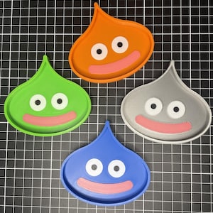 May include: Four colorful, droplet-shaped trays in orange, green, blue, and gray. Each tray features two white circles for eyes and a pink, curved mouth. The trays are arranged on a black grid background.