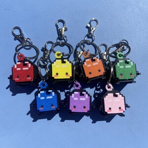 May include: A collection of pixelated bug keychains in various colors, including red, yellow, orange, green, blue, purple, and pink. Each keychain features a silver clasp and ring for attaching to keys or bags. The bugs have a cute, smiling face.