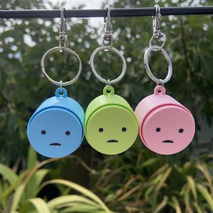 May include: Three keyrings in blue, green, and pink. Each has a cylindrical charm with a small face and a silver ring. The keyrings are suspended against a blurred green backdrop, with a focus on the colorful charms.