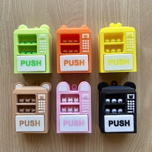 May include: Six colourful miniature vending machine figurines. Each has a "PUSH" button and a small display of tiny figures. Colours include green, orange, yellow, brown, pink, and black. The vending machines have bear-shaped ears.