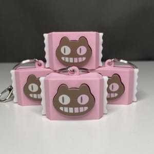 May include: A collection of pink and brown cat-faced keyrings. Each keyring has a brown cat face with oval eyes and a smiling mouth on a pink rectangular base. They have a white, ruffled border and a silver keyring.