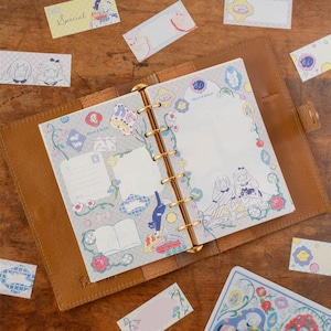Könnte beinhalten: Ein offener brauner Lederordner mit goldenen Ringen, umgeben von dekorativen Aufklebern und einer Notizbuchseite. Die Notizbuchseite hat einen floralen Rand und Illustrationen. Einige Aufkleber tragen das Wort "Special".