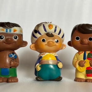 May include: Three vintage figurines depicting diverse characters. The figures have brown skin and are dressed in colorful outfits. One figure holds a small weapon, another wears a turban, and the third holds a small guitar. The figurines are on a white surface.