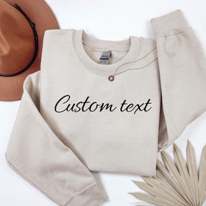 May include: A beige sweatshirt with the words "Custom text" in black script. A gold necklace with a pendant is on the sweatshirt. A brown hat and a dried palm leaf are in the background.