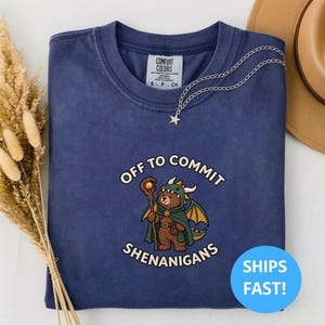 Puede incluir: Una camiseta azul marino de cuello redondo con un gráfico de un oso pardo vestido de dragón con un bastón, rodeado por el texto "OFF TO COMMIT SHENANIGANS". La camiseta tiene una etiqueta Comfort Colors en el cuello y un collar de estrella plateado.
