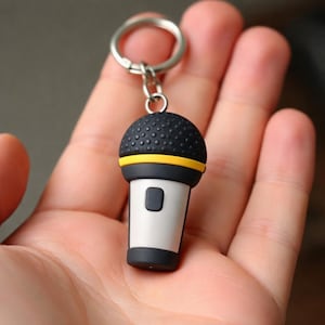 May include: A novelty microphone keychain. The keychain features a black, yellow, and white microphone design. The microphone has a black top with small bumps, a yellow band, and a white body with a black button. A silver keyring is attached.