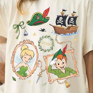 Könnte beinhalten: Elfenbeinfarbenes T-Shirt mit Cartoon-Illustrationen von Peter Pan-Charakteren und einem Piratenschiff. Das Design umfasst Tinkerbell, Peter Pan, einen Piratenhut und das Wort "Neverland" in einem Kranz.