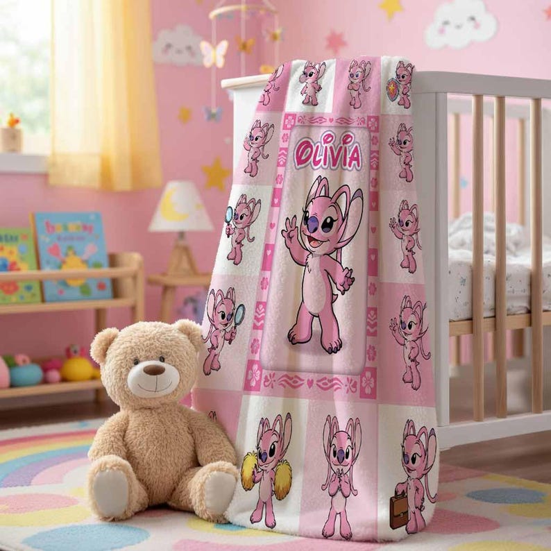 Op de afbeelding: Een roze en witte babydeken met een cartoonkarakter en de naam "Olivia". De deken is over een witte wieg gedrapeerd en heeft een herhalend patroon van het karakter. Een knuffelbeer zit voor de wieg.