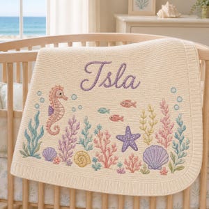 Könnte beinhalten: Eine cremefarbene Strickdecke für Babys mit einer gestickten Unterwasserszene, die ein Seepferdchen, einen Seestern, Muscheln und Seetang in Pastelltönen zeigt. Der Name Isla ist mittig in violetter Schreibschrift eingestickt.