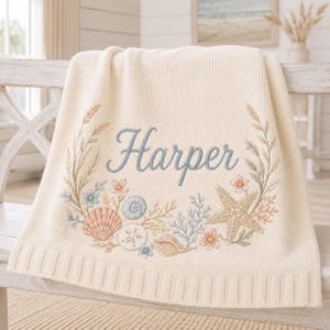 Peut inclure: Une couverture en tricot de couleur crème ornée d'une broderie représentant une étoile de mer, divers coquillages et des brins de fleurs disposés en couronne. Le prénom Harper est brodé en lettres cursives bleues au centre.