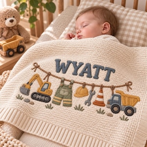 Könnte beinhalten: Eine cremefarbene Strickdecke für Babys mit einem gestickten Baustellenmotiv und dem Namen WYATT in dunkelblauen Buchstaben. Das Design zeigt einen gelben Bagger, eine grüne Latzhose, einen gelben Bauhelm, einen Schraubenschlüssel, eine Pylone und einen Kipper.
