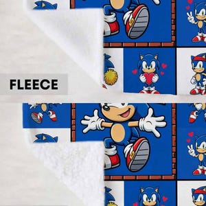 May include: A blanket featuring a grid pattern of Sonic the Hedgehog in various poses, including jumping and holding a red heart. The top section is labeled FLEECE with a smooth texture, and the bottom section is labeled SHERPA with a textured surface.