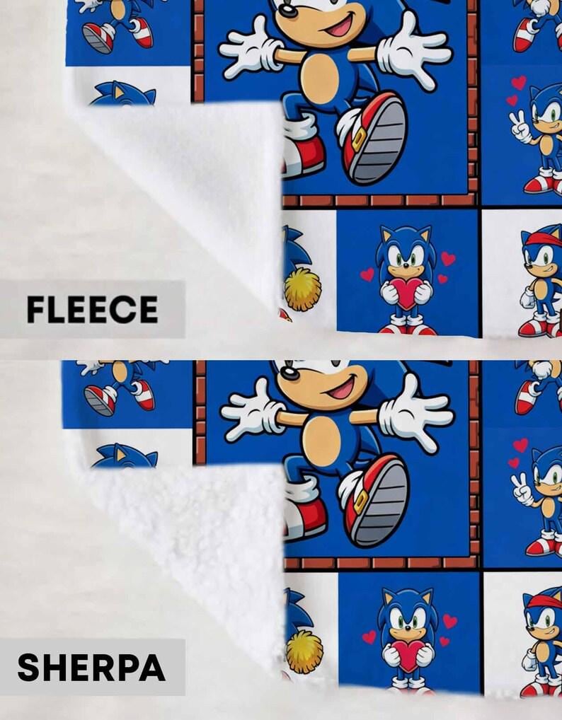 May include: A blanket featuring a grid pattern of Sonic the Hedgehog in various poses, including jumping and holding a red heart. The top section is labeled FLEECE with a smooth texture, and the bottom section is labeled SHERPA with a textured surface.