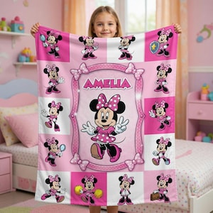 Może przedstawiać: R&oacute;żowy polarowy koc z Minnie Mouse w r&oacute;żnych pozach. Koc ma centralną ramkę z napisem "AMELIA" i jest ozdobiony kokardkami. Myszka Minnie ma na sobie r&oacute;żową sukienkę w kropki i kokardę.