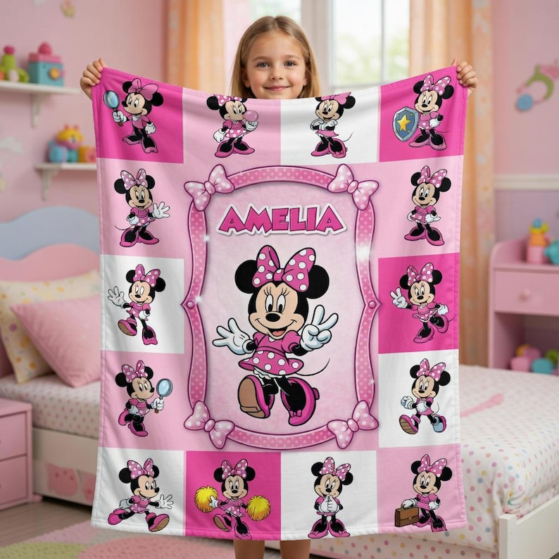 Może przedstawiać: R&oacute;żowy polarowy koc z Minnie Mouse w r&oacute;żnych pozach. Koc ma centralną ramkę z napisem "AMELIA" i jest ozdobiony kokardkami. Myszka Minnie ma na sobie r&oacute;żową sukienkę w kropki i kokardę.