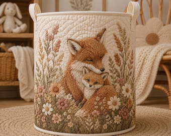 May include: A cylindrical cream-colored fabric storage basket featuring a quilted texture and an embroidered design of an adult fox and a kit nestled together among tall grasses and various flowers in shades of orange, pink, and white.