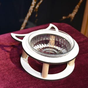 Könnte beinhalten: Ein weißer, katzenförmiger Futternapfständer mit einer durchsichtigen Glasschale. Der Ständer hat eine kreisförmige Basis und wird von drei Holzbeinen getragen. Die Schale ist für eine Katze oder ein kleines Haustier konzipiert.