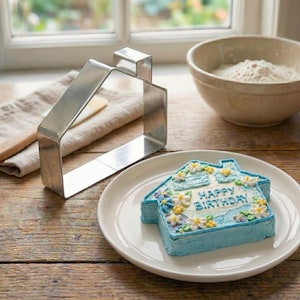 May include: A stainless steel house-shaped cookie cutter next to a small blue frosted cake decorated with white and yellow flowers. The cake says "Happy Birthday" in blue frosting on a white plate.