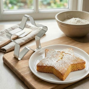 May include: A star-shaped cake dusted with powdered sugar sits on a white plate. A stainless steel star-shaped cookie cutter is next to the cake. A wooden cutting board, a bowl of flour, and a kitchen towel are in the background.