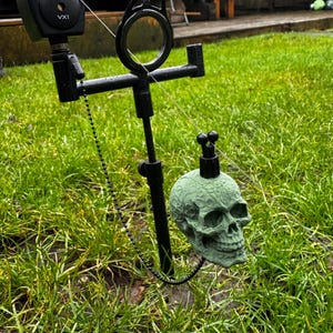 May include: A black fishing rod rest with a green skull-shaped decorative element. The skull is suspended from a black chain and rod. The background is a green lawn.