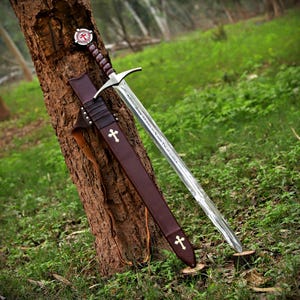 May include: A steel sword with a silver blade and a dark brown leather-wrapped hilt featuring a circular pommel with a red cross emblem. The sword is accompanied by a matching dark brown leather scabbard adorned with two small white crosses.