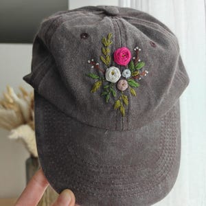 Peut inclure: Casquette de baseball marron avec un motif floral brodé. La broderie présente une rose rose, des fleurs blanches et bleu clair, et des feuilles vertes. La casquette est en tissu doux et texturé.