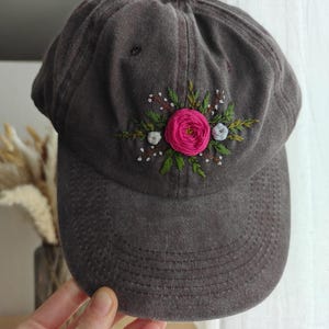 Peut inclure: Casquette de baseball gris foncé ornée d'une broderie florale. La broderie présente une grande rose rose, de petites fleurs blanches et des feuilles vertes. La casquette a une visière incurvée et un bouton sur le dessus.