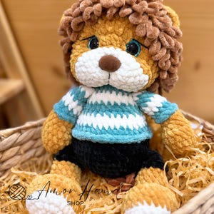 Puede incluir: Un juguete de león de peluche hecho a mano con una melena marrón, una nariz marrón claro y ojos azules. El león lleva un suéter a rayas azules y blancas y pantalones cortos negros. El juguete está sentado en una cesta tejida con paja.