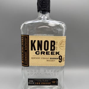 May include: An empty clear glass bottle of Knob Creek Kentucky Straight Bourbon Whiskey. The label features black text on a tan background, stating Aged Nine Years, 100 Proof, and 50% Alc/Vol. The bottle has a black wax seal on the cap.