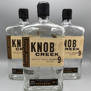 May include: Three clear glass bottles of Knob Creek Kentucky Straight Bourbon Whiskey. Each bottle features a tan label with black text stating Aged Nine Years, 100 Proof, and Small Batch. The bottles have black caps and embossed glass details.