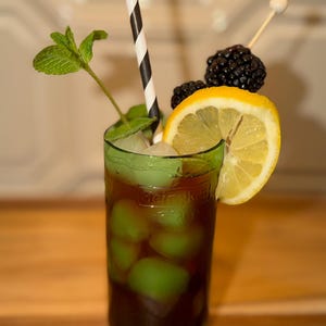 May include: A dark-colored cocktail in a green glass with the embossed text Heineken. The drink contains light green ice cubes, a black and white striped paper straw, a sprig of fresh green mint, a lemon slice, and two blackberries on a skewer.