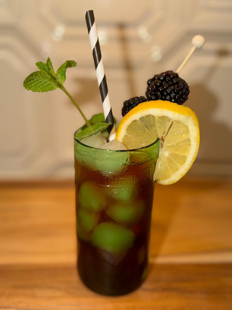 May include: A dark-colored cocktail in a green glass with the embossed text Heineken. The drink contains light green ice cubes, a black and white striped paper straw, a sprig of fresh green mint, a lemon slice, and two blackberries on a skewer.