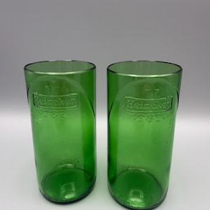 May include: Two cylindrical drinking glasses crafted from green glass bottles. Each glass features the embossed Heineken logo and a star symbol on the front surface. The glasses have a smooth, straight-sided design with a clear, open top rim.